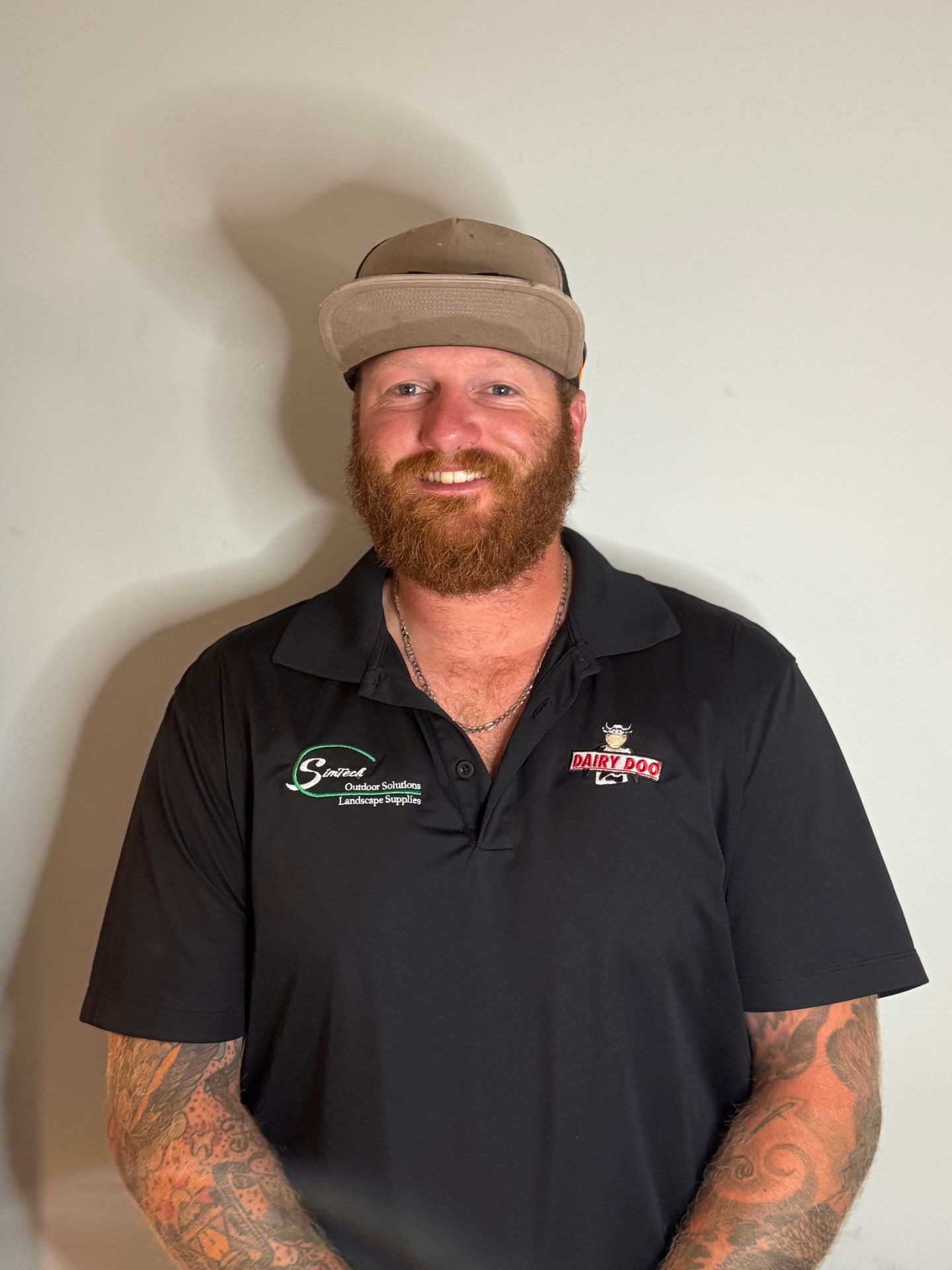 Bearded man in a black polo shirt and hat smiling against a plain background.