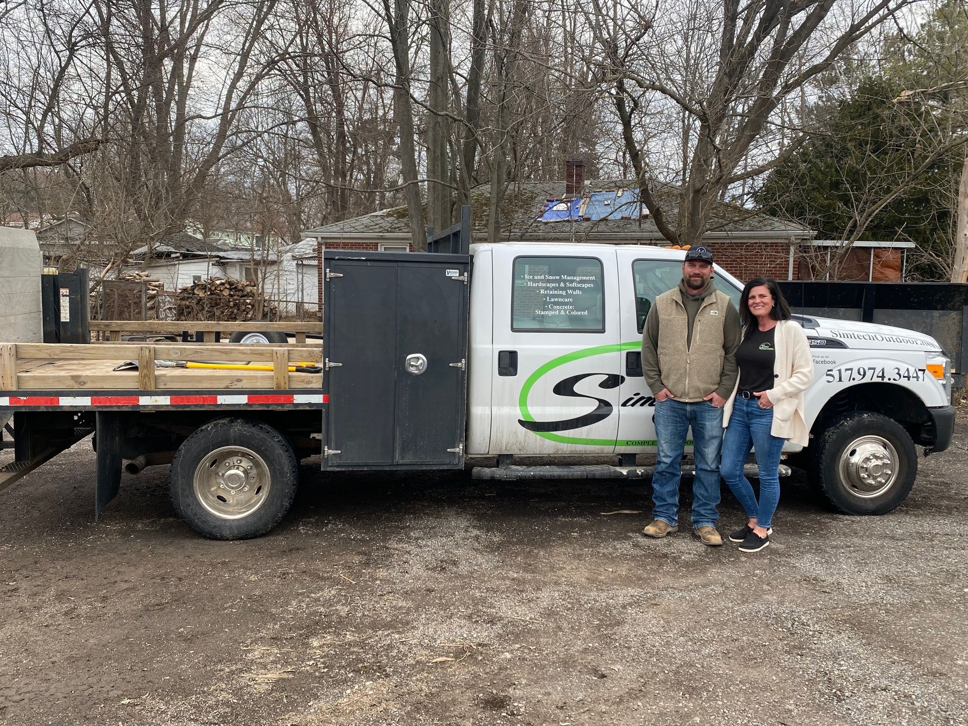 Two people standing in front of a work truck parked outdoors near trees and buildings.